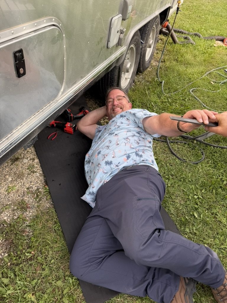 A man lying on a mat under an Airstream, smiling up at the camera, cordless drill and pliers beside him