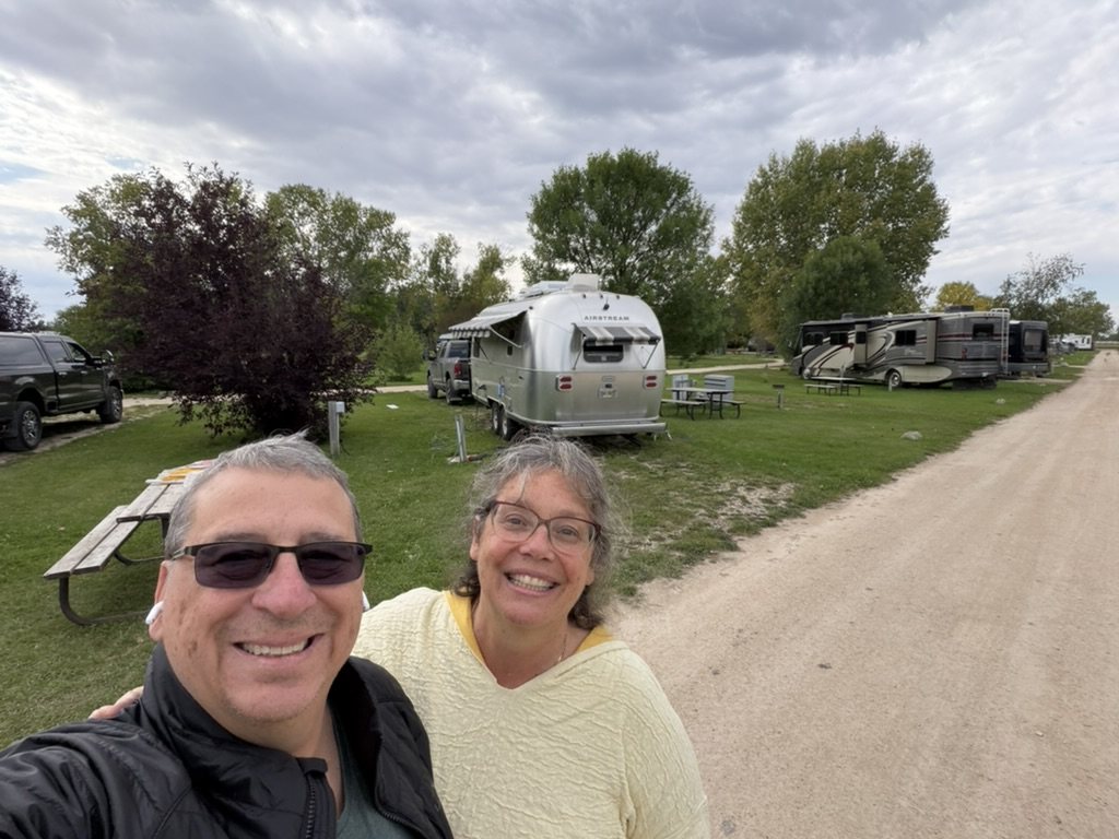 Two of us in front of an Airstream and tow truck at a grassy campsite, other rigs down the road