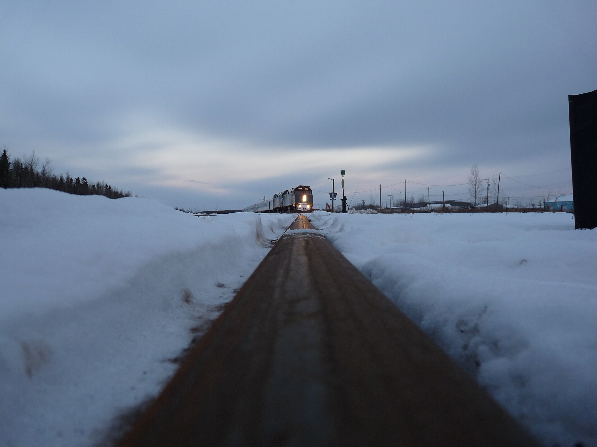 A Hudson Bay Line train approaching along a snow-banked single track at Thompson, Manitoba, headlights on under a grey sky.