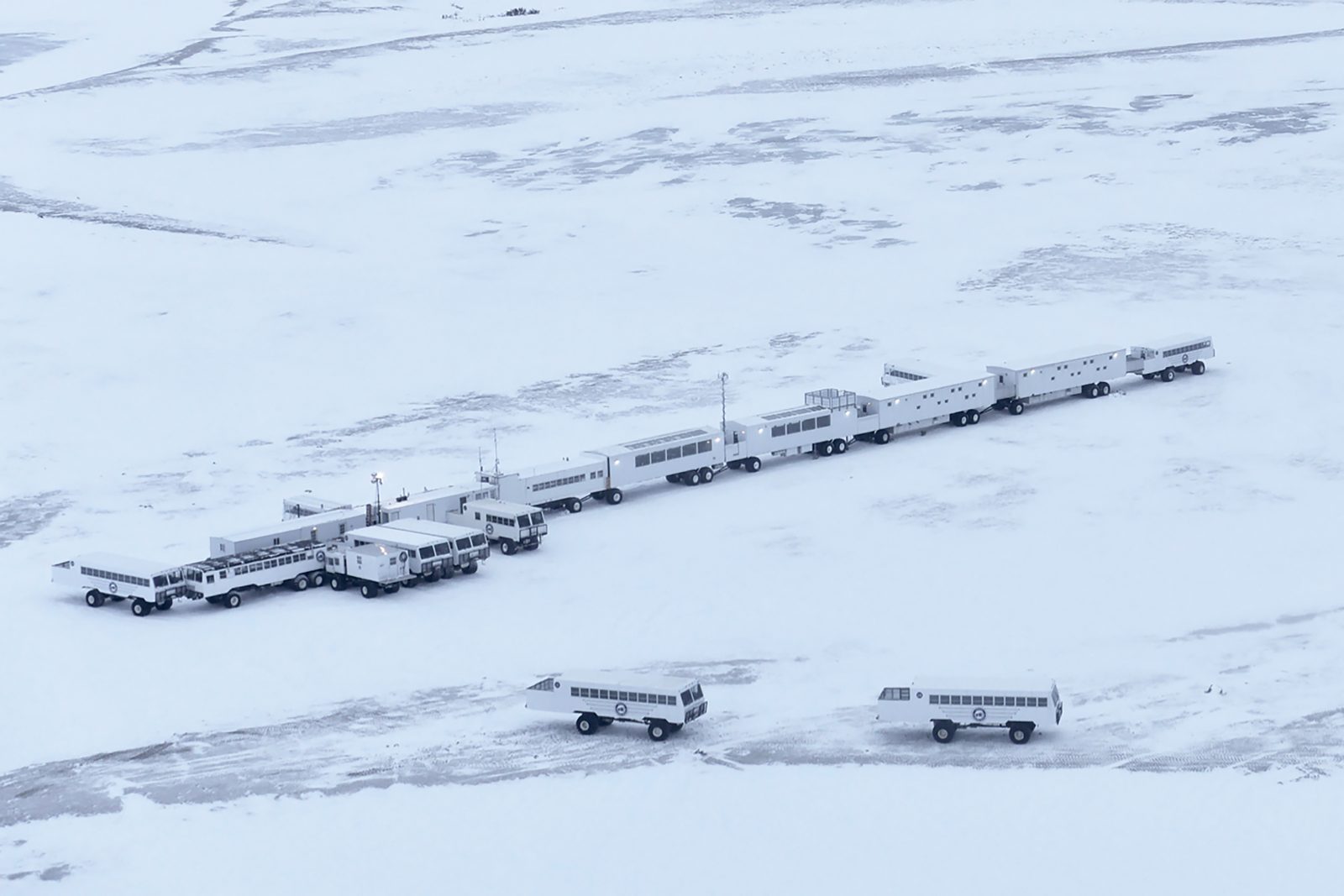 Aerial view of the Tundra Buggy Lodge complex on snow-covered tundra — a long line of linked white units with several Tundra Buggies parked nearby.