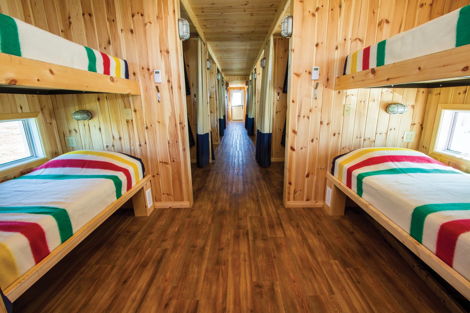 The interior corridor of a Tundra Buggy Lodge sleeper unit: pine-panelled walls, curtained berths on both sides, upper and lower bunks made up with striped Hudson's Bay point blankets.