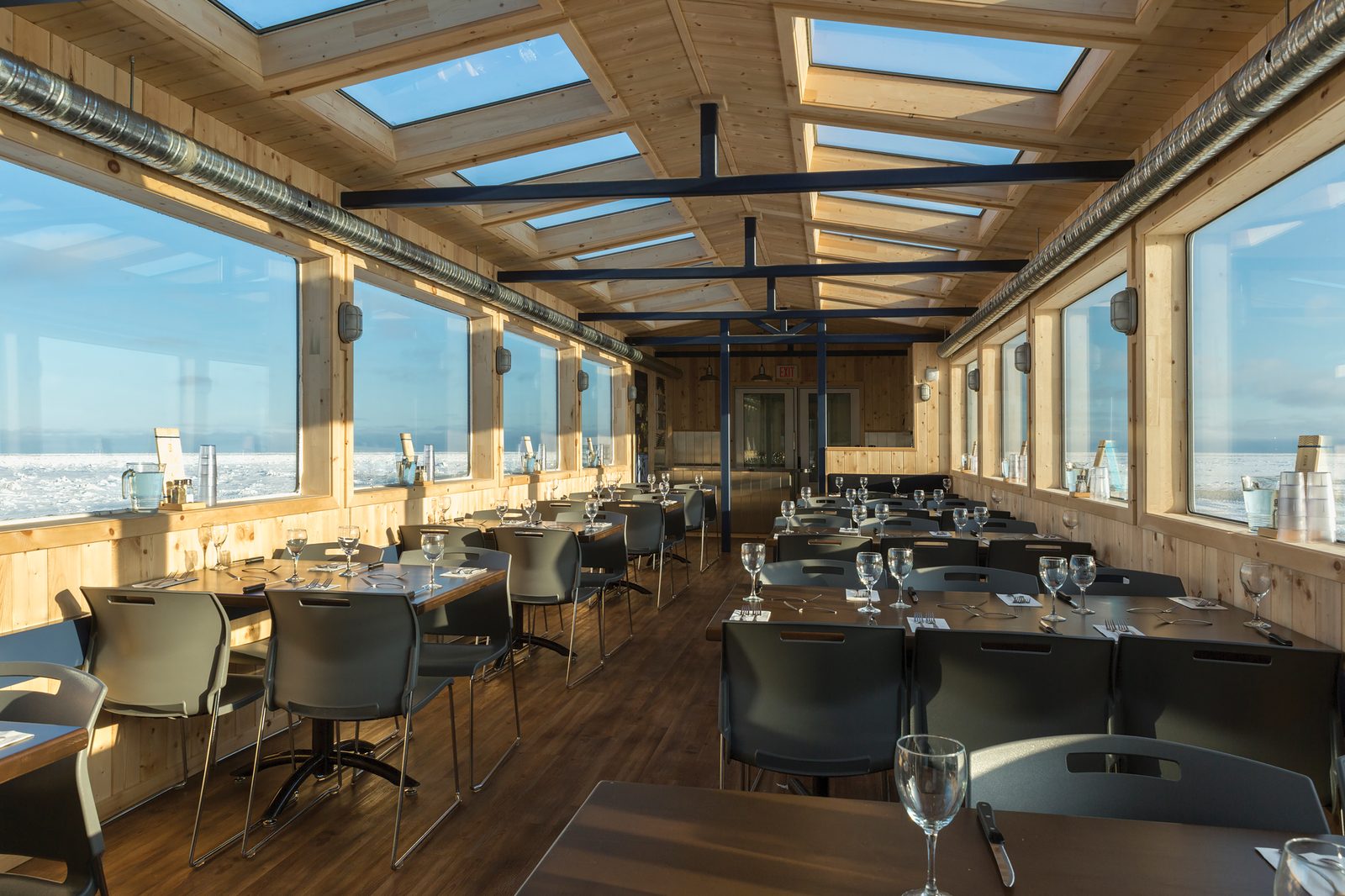 The Tundra Buggy Lodge dining unit: long pine-lined room with skylights overhead, tables set with glassware, and sea ice visible through panoramic windows on both sides.