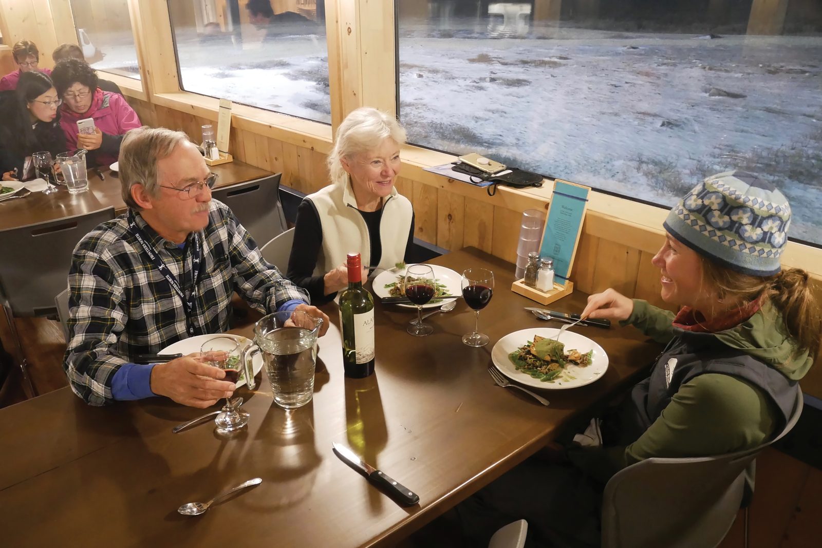 Three guests seated at a dinner table in the Lodge with wine and plated food, frost-covered tundra visible through the window beside them.